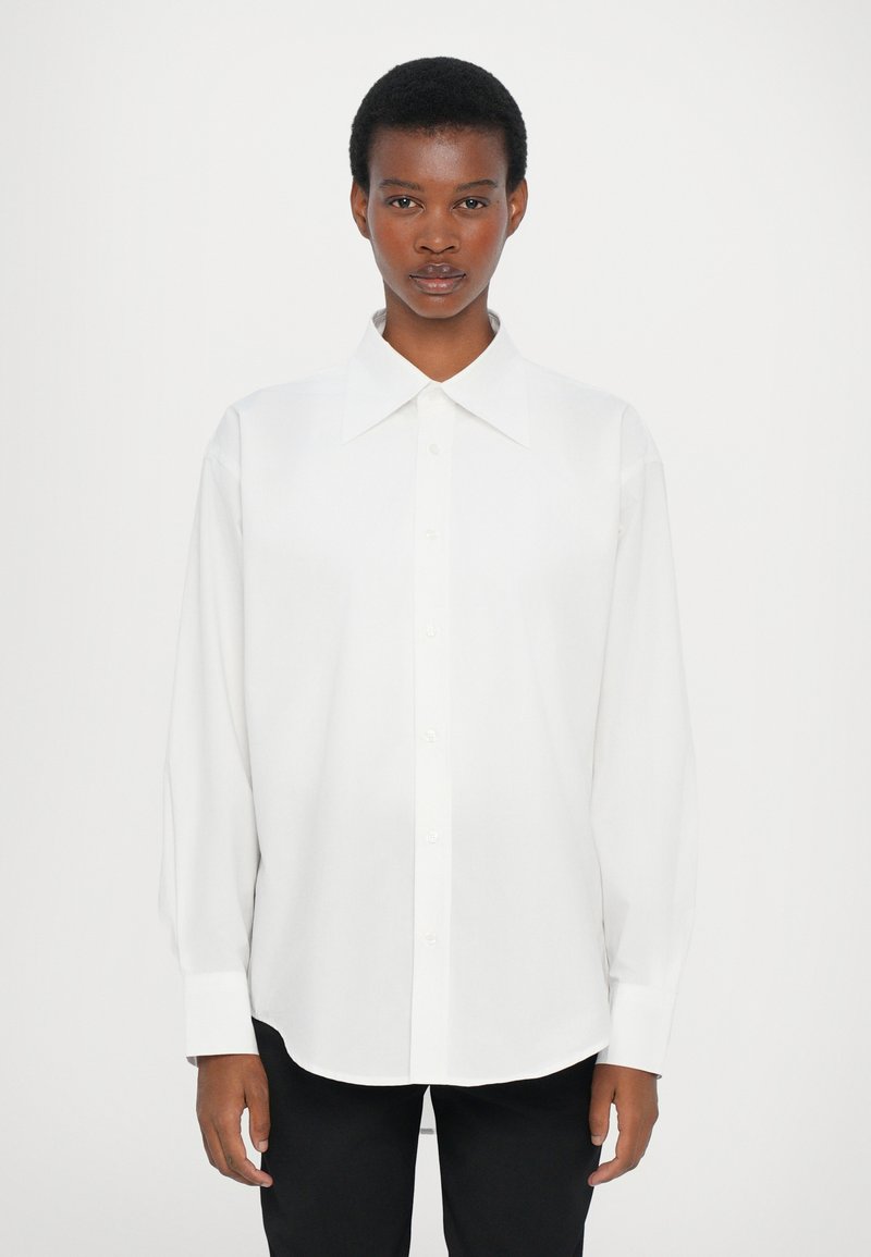 White long-sleeve button-up shirt with a large pointed collar, made of smooth fabric. Features a straight hem and loose fit.