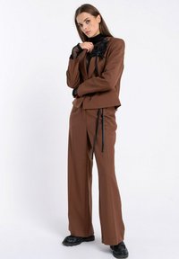 Brąsłowy garnitur o dopasowanym kroju z szerokimi spodniami i krótką marynarką. Uzupełniony czarnym kwiatowym detalem na ramieniu i czarnym topem pod spodem.