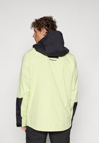 Veste imperméable jaune clair avec une capuche noire contrastante, présentant un logo dans le dos et des accents noirs sur les manches.