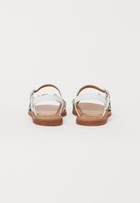 Sandales blanches avec des embellissements floraux sur les brides, une semelle plate marron et des boucles réglables pour un ajustement sécurisé.