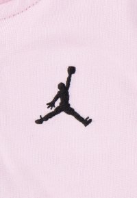 Logo Jumpman noir sur tissu rose pâle, mettant en avant une silhouette athlétique avec des membres étendus et un ballon de basket, des coutures détaillées visibles.