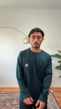 Tamsiai žalia, ilgomis rankovėmis marškinėliai iš minkštos medžiagos, su nedideliu logotipu ant krūtinės. Modelis stovi susikabinęs rankas, apsuptas vidaus dekoracijų.