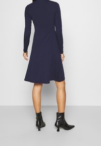 Granatowa sukienka z długim rękawem, o dopasowanym torsie i rozkloszowanej spódnicy, wykonana z miękkiego materiału. Noszona z czarnymi botkami za kostkę z ozdobnym obcasem.
