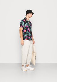 Schwarzes Kurzarmhemd mit buntem Blumen- und Palmenblattmuster, kombiniert mit hellbeigen Hosen und weißen Sneakern, während eine beige Tragetasche gehalten wird.