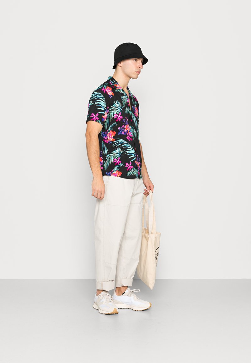 Schwarzes Kurzarmhemd mit buntem Blumen- und Palmenblattmuster, kombiniert mit hellbeigen Hosen und weißen Sneakern, während eine beige Tragetasche gehalten wird.