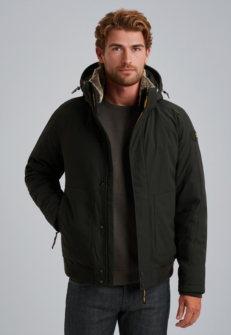 Giacca nera isolante con cappuccio foderato in pelliccia, chiusura con zip e bottoni a pressione, due tasche frontali e polsini a costine. Indossata sopra un maglione grigio.