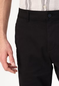 Černé kalhoty s hladkou texturou, vybavené přední kapsou a zapínáním na knoflík. Podrobné švy a štíhlý střih.