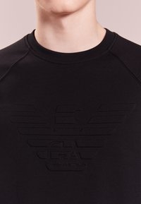 Černá mikina s kulatým výstřihem a gumovým logem orla uprostřed. Má hladkou texturu a raglánové rukávy.