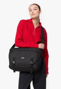 Borsa a tracolla nera con tracolla regolabile, chiusura con fibbia e finitura texturizzata. Presenta una patch con il logo e un design minimalista.