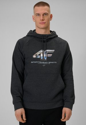Mężczyzna w ciemnoszarej bluzie z kapturem z srebrnym logo "4F Sportswear Brand", zwrócony przodem na jednolitym jasnoszarym tle.