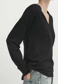 Personne portant un pull noir à col en V à manches longues et un jean bleu clair, la main dans la poche, vue du cou aux hanches.