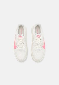 Pár bílých tenisek Nike Air Force 1 s růžovými logy ve tvaru háčku, bílými tkaničkami a růžovým označením na jazyku, pohled zhora.