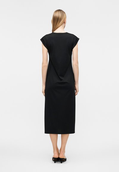 Γυναίκα με ίσια ξανθά μαλλιά που φοράει αμάνικο μαύρο φόρεμα μέσης και μαύρα παπούτσια με τακούνι, στέκεται με την πλάτη προς τα εμπρός σε απλό φωτεινό φόντο.