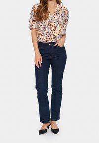 Blouse florale à manches courtes dans des tons beige, bleu et marron ; jean droit bleu marine ; escarpins noirs à bout pointu.