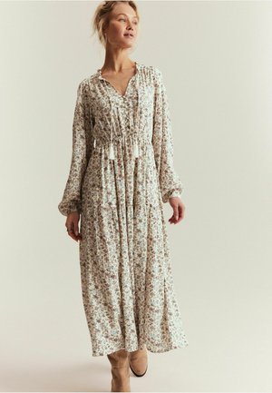 Kvinde iført en lang blomsterkjole med lange ærmer og frynset snøre, stående foran en ensartet lys baggrund.