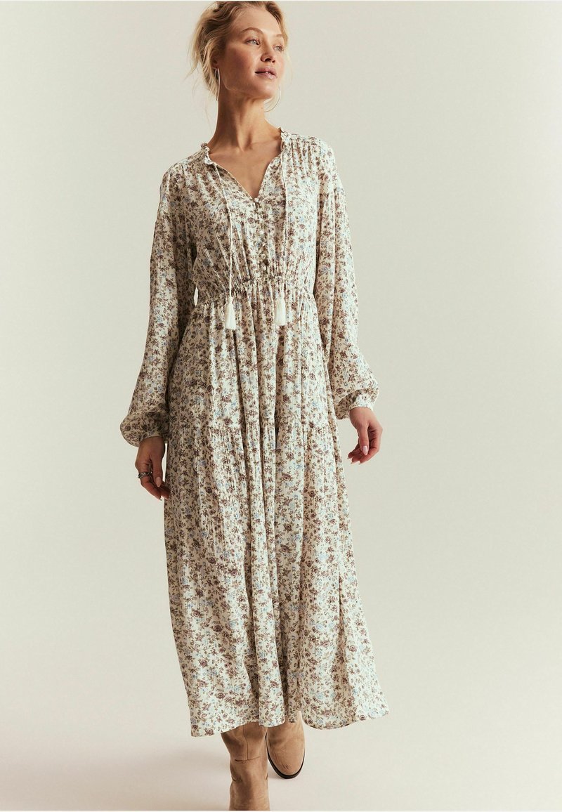 Frau in einem langen, floralen Kleid mit langen Ärmeln und Quastenbändern, stehend vor einem schlichten, hellen Hintergrund.