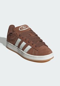 Brown suede sneaker featuring white three stripes on the side, round logo on the tongue, and a gum rubber outsole.