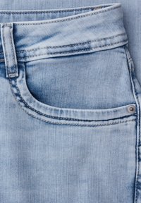 Gros plan sur un jean en denim bleu clair montrant la poche avant, la boucle de ceinture et le détail des rivets en métal sur la texture du tissu.