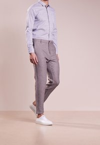 Man med mönstrad skjorta med knappar, grå smala byxor och vita sneakers som står på ett beige golv mot en enfärgad bakgrund.