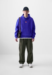Felpa viola con logo, pantaloni cargo olive, t-shirt bianca sotto, cappellino nero e sneakers grigie; porta una piccola borsa a tracolla.
