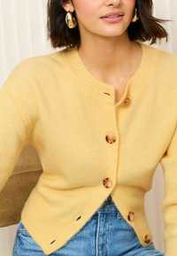 Cardigan jaune en matériau doux, avec un col rond, cinq grands boutons et une coupe légèrement courte et ajustée.