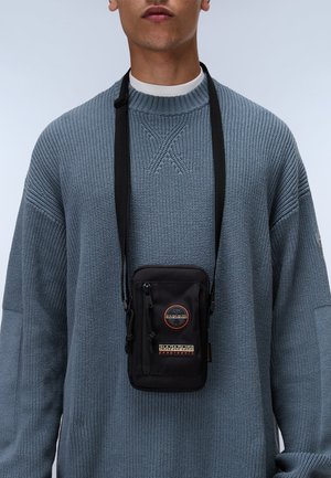 Black crossbody bag with front zip pocket, logo patch, and adjustable strap. Worn over a light blue textured sweater with a crew neckline.
