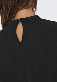 Haut noir avec un col haut et une ouverture en forme de clé à l'arrière, fabriqué en tissu texturé avec un aspect légèrement froissé.