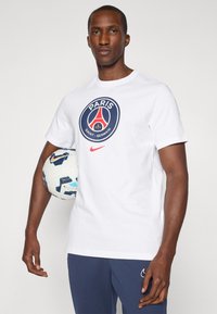 Hvid bomulds t-shirt med et blåt og rødt Paris Saint-Germain logo, parret med marineblå bukser. Figuren holder en mønstrede fodbold.
