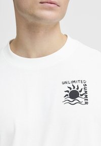 Camiseta de algodón blanca con un gráfico negro de un sol y olas, con el texto "VERANO ILIMITADO" posicionado en el pecho.