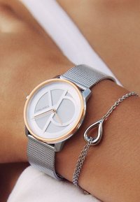 Sidabrinis tinklinis laikrodis su baltu ir šaltiu dialu, kuriame yra geometriniai akcentai. Papildytas sidabriniu apyranke su ašarėlės formos pakabuku.