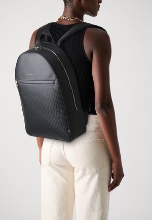 Black backpack with a textured front, smooth sides, and a top handle. Features a zipper closure and subtle logo detail. Worn on the back.