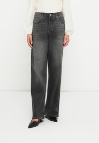 Jeans wide-leg in denim grigio scuro con tasche anteriori, passanti per cintura e un leggero effetto sbiadito. Abbinati a un top chiaro e strutturato e tacchi a punta neri.