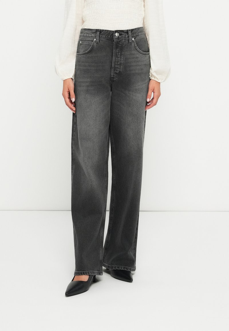 Jeans wide-leg in denim grigio scuro con tasche anteriori, passanti per cintura e un leggero effetto sbiadito. Abbinati a un top chiaro e strutturato e tacchi a punta neri.