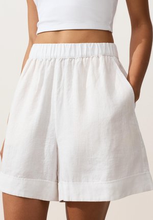 Nő magasderekú fehér len rövidnadrágot visel rugalmas derékpánttal és oldalzsebekkel, fehér rövidített top társaságában.