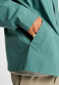 Giacca maglia color verde petrolio con vestibilità morbida, dotata di una tasca laterale e realizzata in tessuto leggero e liscio. Indossata sopra una camicia grigio chiaro.