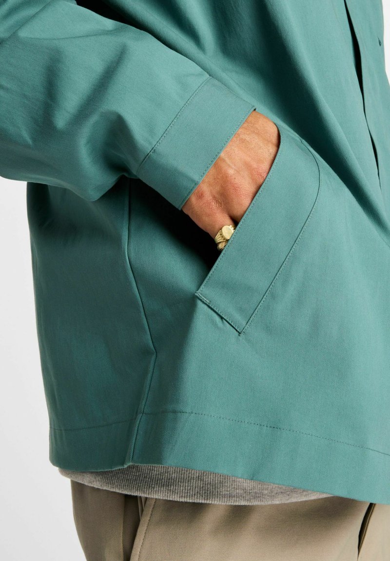 Türkisfarbene Hemdjacke mit lockerem Schnitt, ausgestattet mit einer Seitentasche und einem glatten, leichten Stoff. Gezeigt als Lagenlook über einem hellgrauen Hemd.