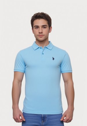 Mladý muž stojí, nosí svetlomodré polo s malým tmavým vyšívaným logom na hrudi a modré džínsy.