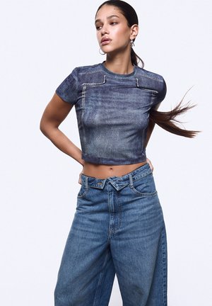 Jeune femme aux longs cheveux foncés porte un top court en denim ajusté à manches courtes et un jean bleu taille haute sur un fond uni.