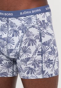 Korte boxershorts i bomull med et blått palmebladmønster på hvit bakgrunn. Livremmen er blå med "BJÖRN BORG" i hvit tekst.