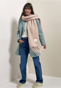 Mintgrön kappa, vit tröja, blå jeans och en texturerad ljusrosa halsduk med fransar. Märkesetikett synlig på halsduken. Avslappnad skor.