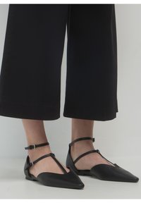 Persona que lleva zapatos planos negros de punta afilada con dobles correas en el tobillo y pantalones anchos negros recortados, contra un fondo liso.