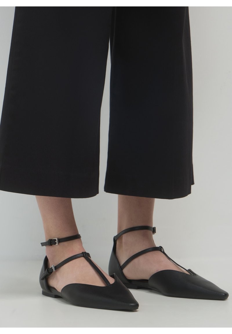 Persona que lleva zapatos planos negros de punta afilada con dobles correas en el tobillo y pantalones anchos negros recortados, contra un fondo liso.