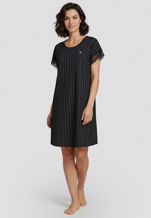 Kvinde stående barfodet iført en sort knælængde kjole med lodrette hvide nålestriber og kortærmer med blonder.