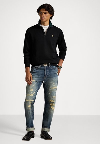 Ung mand iført sort kvart-zip sweater, hvid skjorte under, slidte blå jeans med opfoldede nederkanter, sorte sko og et armbåndsur, stående.