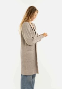 Vrouw met lang haar in een beige gebreid vest en blauwe jeans, die de mouw aanpast op een witte achtergrond.