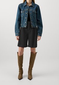 Femme portant une veste en jean courte, un short noir taille haute et des bottes en cuir marron montant jusqu'aux genoux, debout devant un fond uni.