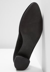 Chaussure à talon haut en daim noir avec semelle en caoutchouc texturée, présentant un bout arrondi et un design minimaliste. Nom de la marque gravé visible.
