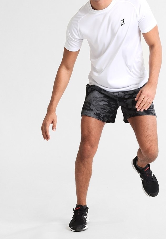 T-shirt de sport blanc à manches courtes avec logo noir, associé à des shorts camouflage noirs et des chaussures de course noires avec semelles blanches.