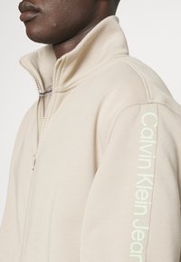 Giacca zip-up beige chiaro realizzata in materiale morbido. Presenta un colletto alto e un logo verticale verde chiaro sulla spalla sinistra.