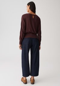 Dunkelburgunderfarbenes Langarmoberteil mit einem runden Schlüssellochdetail im Rücken, kombiniert mit weiten, dunkelblauen Jeans und braunen Holzclogs. Weiche Stofftextur.